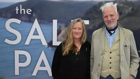Getty Images Two people stood in front of a large screen which has a coastal scene and the words The Salt Path in big white letters. On the left is a woman with long blonde hair, wearing all black, and beside her is a man with spiky white hair wearing a beige jacket and waistcoat, blue shirt and navy cravat. They ar eboth looking at the camera and smiling.