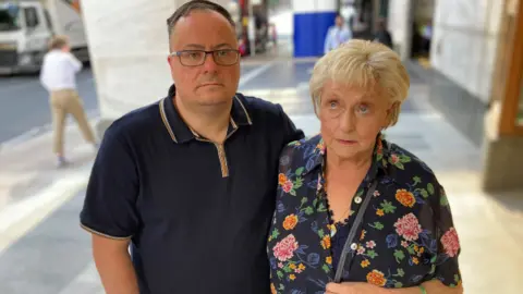 A man in his 40s and his mother stand next to each other. He is wearing glasses and has short dark hair, she is wearing a floral shirt and has blond hair