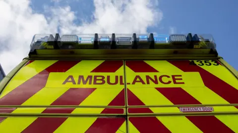 Photo of the back of an ambulance which is yellow and red striped with the word "ambulance" on. 