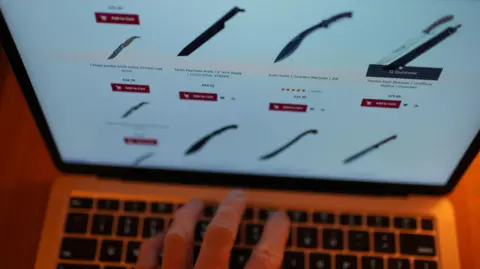 A hand on a laptop keyboard, with a screen showing a display of large knives for sale