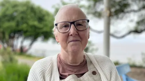 Sally is looking directly at the camera with a neutral expression on her face. She is wearing a cream cardigan, brown t-shirt and light brown dungarees. She has short white hair and glasses.