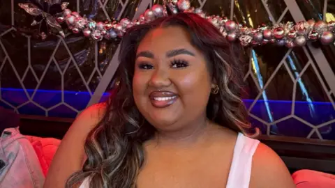 Miah Carter Miah is looking at the camera, wearing full makeup and a white vest top, her long hair is loose. She is sitting in front of a window with a grey, geometric design and a garland of baubles.