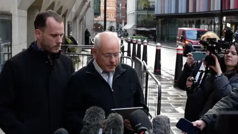 John Fairhall/BBC Steven Hall and Graham Hall standing outside the Old Bailey in front of a press pack. They are both looking downwards at a tablet from which Graham is reading out a statement. 
