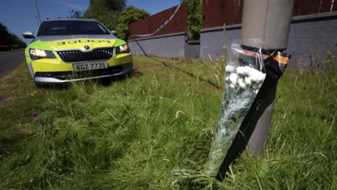A bunch of white flowers leans against a lamppost on a grass verge. A police car and police tape are also visible in the picture