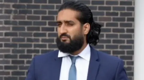 Abubakr Ben Yusaf, who wears a navy two piece suit, a blue tie and white shirt. He has brown long hair and a brown long haired beard. He is leaving Mold Crown Court and holds a blue folder under his left arm