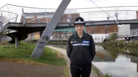 YouTube Khizra Bano standing next to a canal in Coventry, wearing her police uniform. A metal footbridge crosses the canal behind her, a grey structure with thin metal railings. One of the supporting spars has graffiti on the side.