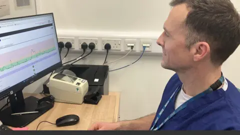 Dr Phil Hammon is sitting by a computer displaying a colourful graph. He is wearing blue scrubs.