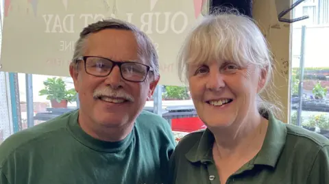 Pauline and Steve Davis, wearing green polo shirts, are stood in the front of their store. There is a window behind them with cars and a street outside.