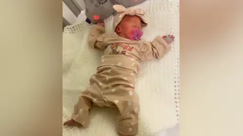 A tiny premature baby lies sleeping in a cot with her arms outstretched. 