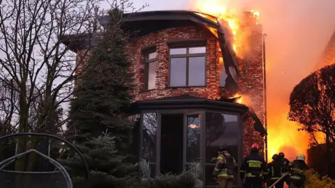 The roof of a modern-looking brick house with large windows burns after being hit by a missile as firefighters in uniform tackle the blaze with a hose