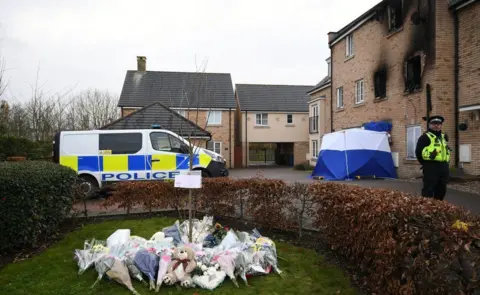 Joe Giddens A modern three-storey terraced house with fire damage around the windows, a police car and officer and piles of flowers, Aynesbury, Cambridgeshire