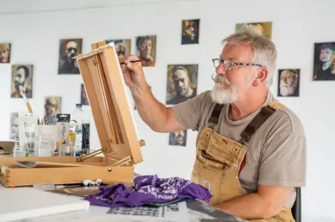 Aaron Law Simon Gee can be seen sitting at an easel painting - with his art exhibited on the walls behind him