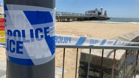 Near the crime scene, an image shows a close-up of police tape wrapped around a metal post and trailing along a metal fence towards the sandy beach. In the background is the sea and Bournemouth Pier.