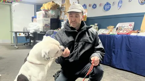 A man wearing a navy raincoat, trousers and grey cap kneeling down next to a white American bulldog which he is holding on a lead. The dog has one paw up on his right arm and he is cupping his hand to give the dog a treat. They are inside a big room with a table behind with a blue tablecloth and various boxes on, there are some decorative blue plates on the wall.