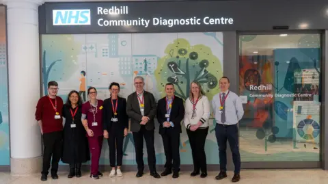 The image shows healthcare workers posing for a photograph outside of Redhill Community Diagnostic Centre. There is a glowing NHS sign and cartoon drawings on the outside of the centre.