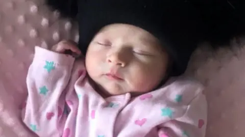 A baby wearing a pink babygrow with blue stars and dark pint hears on it. She is asleep while lying on a pink blanket and wearing a black hat