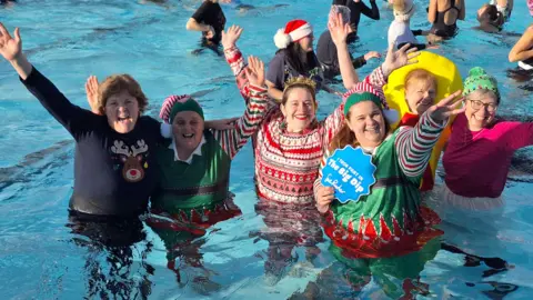 Nic Moorey Six women are pictured standing in an outdoor pool wearing Christmas clothing. One is dressed as an elf, another in a Christmas jumper with Christmas tree earrings. They are standing and smiling with their arms in the air. 