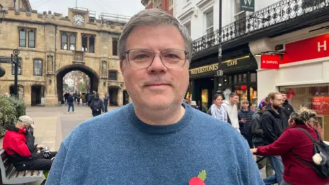 Richard Storey is standing on the High Street in Lincoln, wearing a blue jumper with a remembrance poppy pinned to it. He has short grey hair and is wearing glasses. The stopped clock is behind him in the distance. It is part of a stone building with an arch.