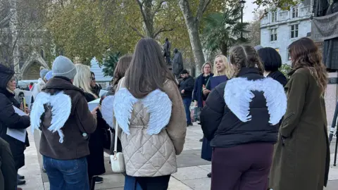 A group of women are stood listening to another women talk. The image is taken from behind and they are all wearing white angel wings. 