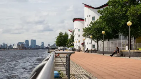 University of East London The River Thames with Canary Wharf in the distance and a path along the river past trees and white, cylindrical buildings that is student accommodation at the Docklands campus of the University of East London