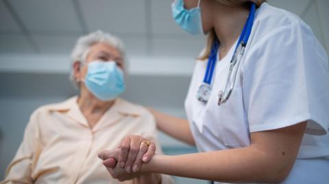 A nurse and elderly woman wearing PPE face masks, holding hands with hospital room in the background