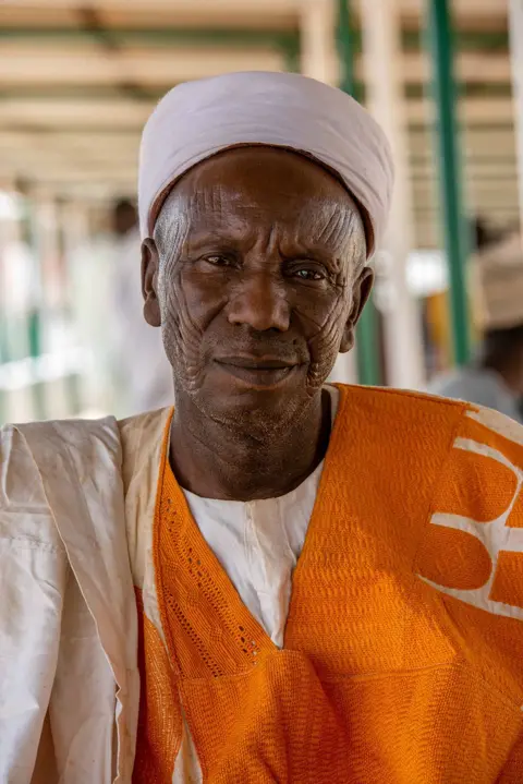 Sani Maikatanga A close-up head-and-shoulders portrait of a man in an orange and white traditional top and white turban.