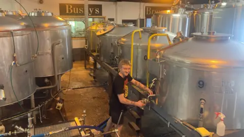 Bushy's Large silver cauldrons lined up in a small room. They have pumps and tubes coming from them. A man in shorts and a T-shirt pours beer from a tap into a silver jug.