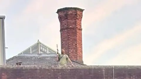 The blurred shape of a man in a green top holding what appears to be a rifle on the roof of a brick building with a large brick chimney behind him