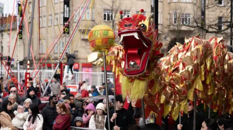 A red Chinese dragon with gold and yellow decorations parades through Sheffield while lots of people watch.