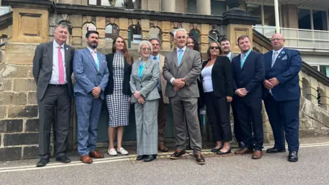 11 people in suits stood infront of grey stone building