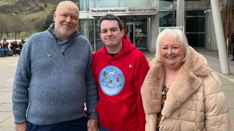 The family smiling outside the Scottish Parliament. Calum, in the middle, is wearing a red jumped with a 'Calum's law' logo 