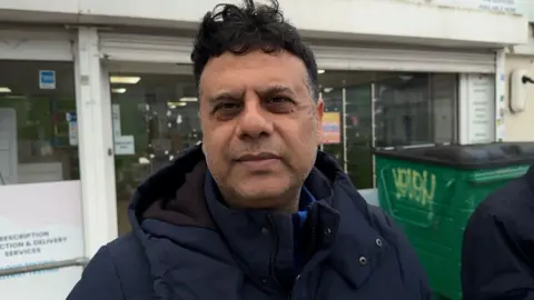 Areef Suhail has short dark hair and is wearing a coat. He is stood outside and glass shop front.