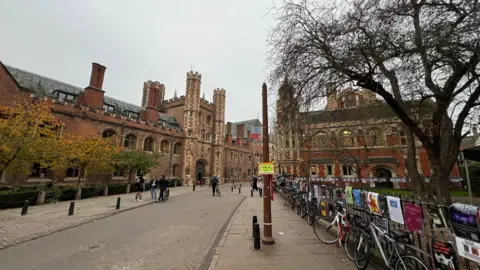 Steve Hubbard/BBC Stock image of Cambridge in the city on a cloudy day. There are bikes against a railing on the right and a grand building on the left. People are standing on the street, which is not busy.