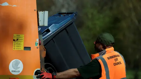 Reuters A man in an orange hi-viz vest which says Birmingham City Council on the back loads a large black bin into an orange waste lorry.