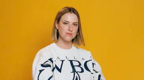 A woman with short blonde hair and earings wearing a white top sitting for a head shot in front of a yellow background.