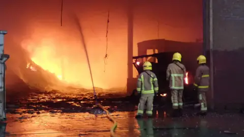 Three fire fighters are standing at the entrance to a warehouse with a large fire taking place inside it. A yellow hose is running along the ground and into the opening to the building, where it is spraying water towards the fire.