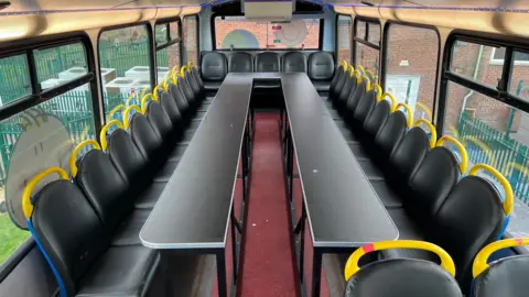 A view of an empty classroom area on the upper deck of the bus. Bus seats have been put around the outside of a long work table.