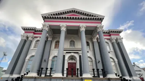 It's a shot of the outside of the Temple. There are eight columns supporting the over-hanging roof. It's painted a dull, grey blue with white sections. The top sections are painted pink. It's a historic looking building. 