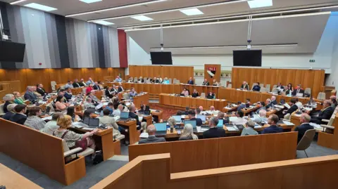 Paul Moseley/BBC A modern council chamber with tiered wooden seating arranged in a semicircle. Numerous attendees sit at desks with laptops and papers. At the front, a raised panel of officials sits beneath a crest, flanked by large screens.