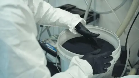 A white bucket containing a black substance with a person in white overalls and black gloves holding the rim