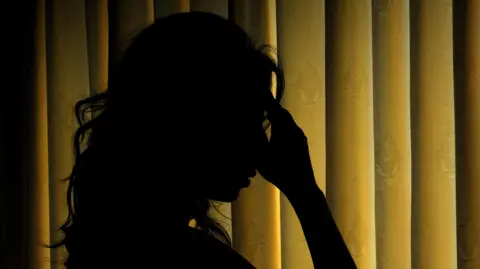 PA Media Long haired woman in silhouhette holding hand to her heads against backdrop of yellow curtains