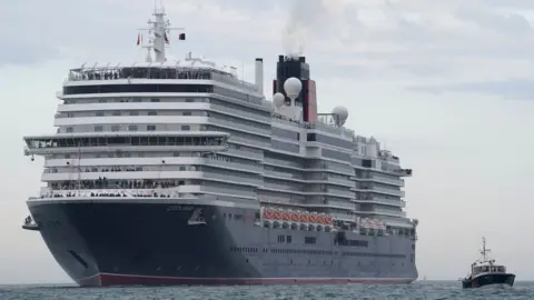 PA Media A large white cruise ship with a navy hull sails through the water. It is being accompanied into a dock by a small boat.