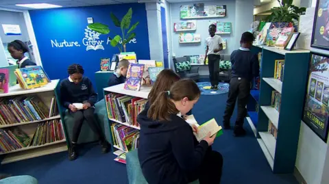 Children sat on benches and in small arm chairs reading books. They are surrounded by book shelves at low level and books on display