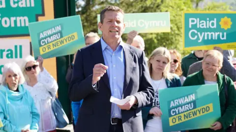 PA Media The image shows Rhun ap Iorwerth standing in front of people holding Plaid Cymru placards. He is wearing a navy suit and is mid-speech.
