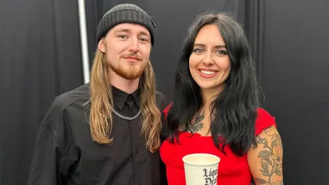 Sean from KNIFE BRIDE is on the left and Mollie is on the right. The two are stood in front of a black curtain and black sofa. Sean is wearing a black beanie and black shirt and Mollie is wearing a red dress and holding a drinks cup.