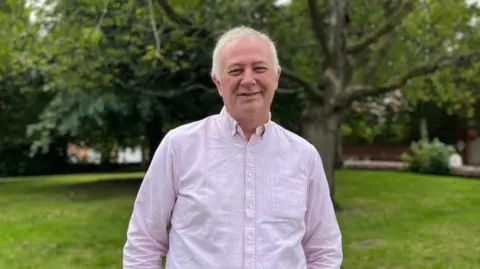 A man with short grey hair stands in a green park smiling into the camera wearing a pink long-sleeved shirt.