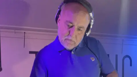 Mark Davis A man with a short grey beard, in a blue polo shirt, with a small logo on it. He has headphones over one ear and is concentrating on his music
