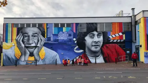 Roopam Carroll A brightly coloured mural on the outside of a building shows Beeston-born Sir Paul Smith wearing a grey jumper and yellow scarf cupping his face with his hands. Next to him is actor Richard Beckinsale wearing a red jacket with black and white film tape behind his head.