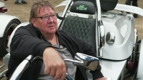A man with dark hair and glasses is sitting in a trike wearing a black zip-up jacket that is open. He has one hand on the handlebar and is looking into the camera.