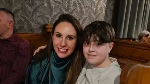 Joanne McLaughlin has dark hair and dark eyes, and is smiling at the camera. She is wearing a turquoise top. Sitting next to her is a young boy with black hair. Behind them is flower wallpaper. 